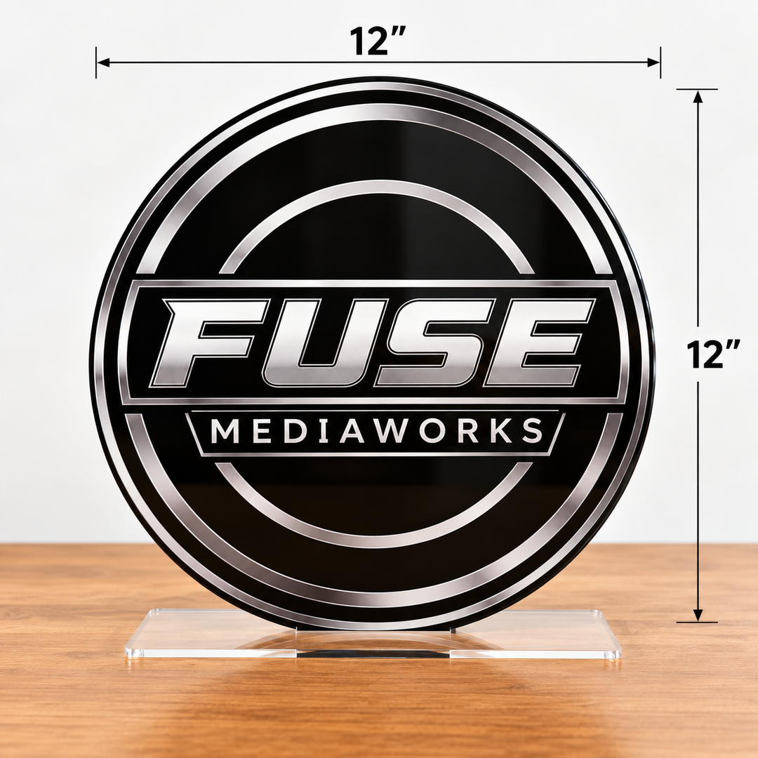 A round black and silver acrylic plaque featuring the 'FUSE MEDIAWORKS' logo, displayed on a clear acrylic stand on a wooden surface. White dimension lines and text indicate the product is 12 inches by 12 inches.