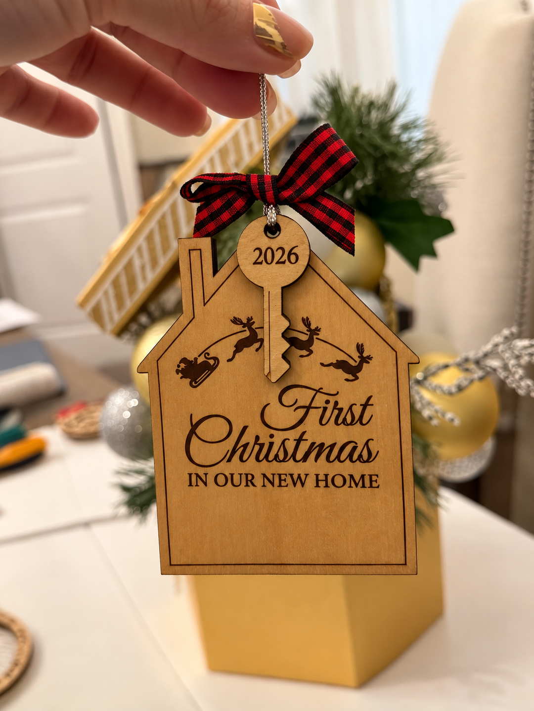 A hand holds a personalized wooden Christmas ornament shaped like a house. The ornament is engraved with &