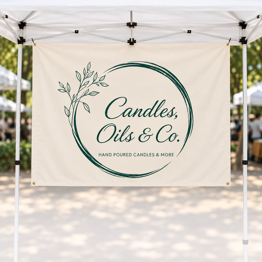 A cream-colored promotional banner hanging from a white pop-up canopy at an outdoor market. The banner features a dark green circular brushstroke design with a leafy branch on the left and the text &