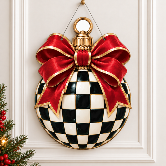 A hanging Christmas ornament shaped like a round ball with a black-and-white checkerboard pattern and a large red satin bow with gold trim, displayed against a white paneled wall with a small Christmas tree visible at the lower left.