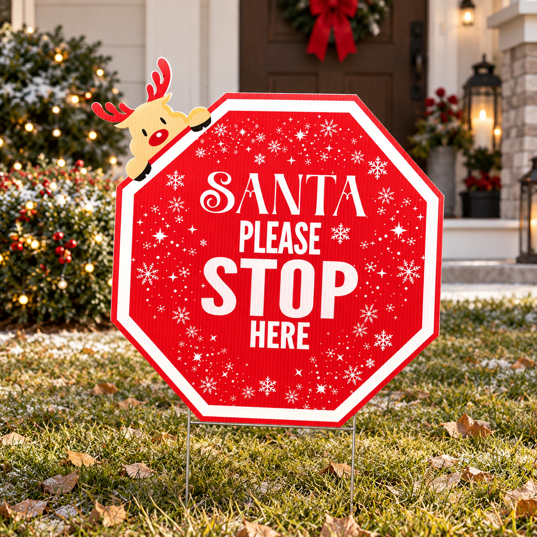 A red holiday yard sign shaped like a stop sign, with white text reading “SANTA PLEASE STOP HERE,” white snowflakes, and a small reindeer peeking over the top edge, displayed on a lawn in front of a decorated house.