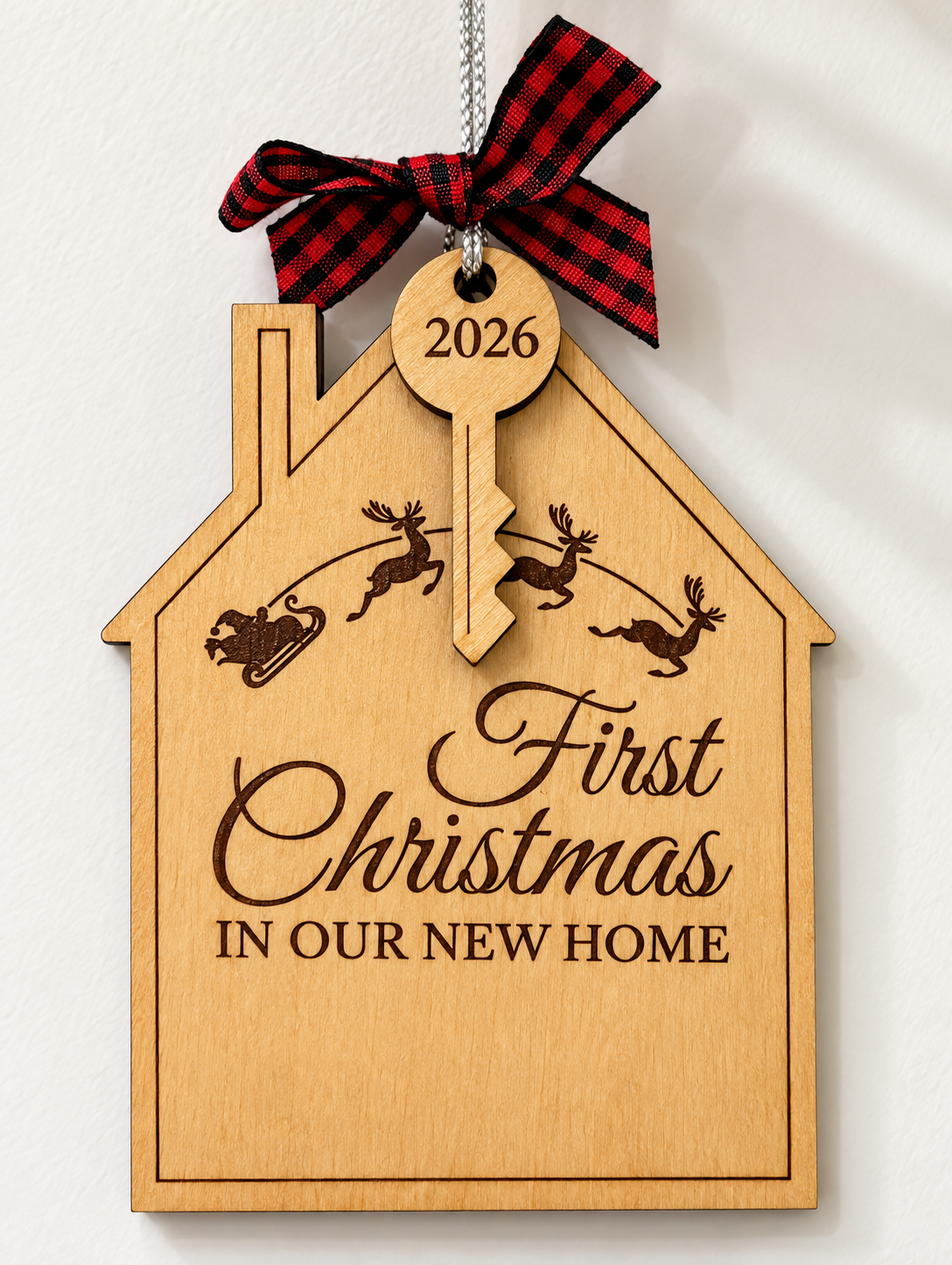 A personalized wooden house-shaped Christmas ornament with a red and black plaid ribbon bow at the top. The ornament is engraved with '2026', 'First Christmas', and 'IN OUR NEW HOME', along with silhouettes of reindeer and a sleigh.
