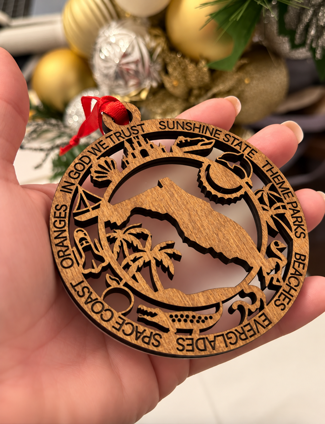 A hand holds a round laser-cut wooden ornament featuring a mountain and beach scene with engraved words like &