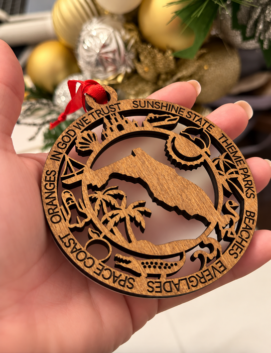 A hand holds a round laser-cut wooden ornament featuring a mountain and beach scene with engraved words like &