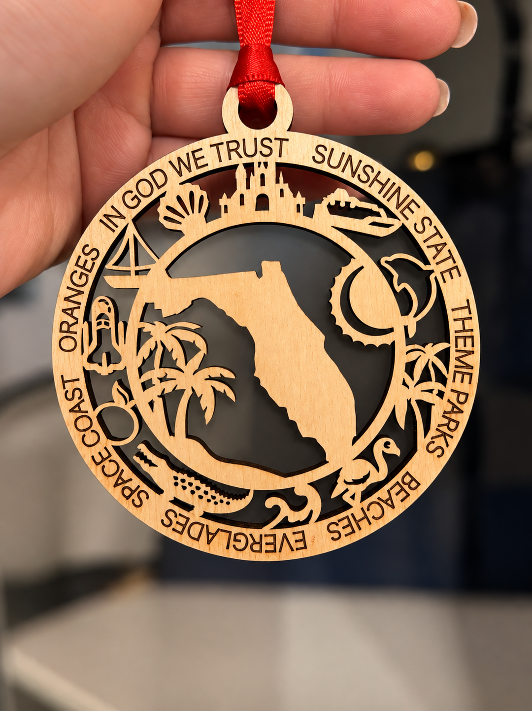 A hand holds a round wooden ornament featuring a cutout Florida state silhouette with surrounding Florida-themed text and icons, hanging from a red ribbon against a blurred dark background.