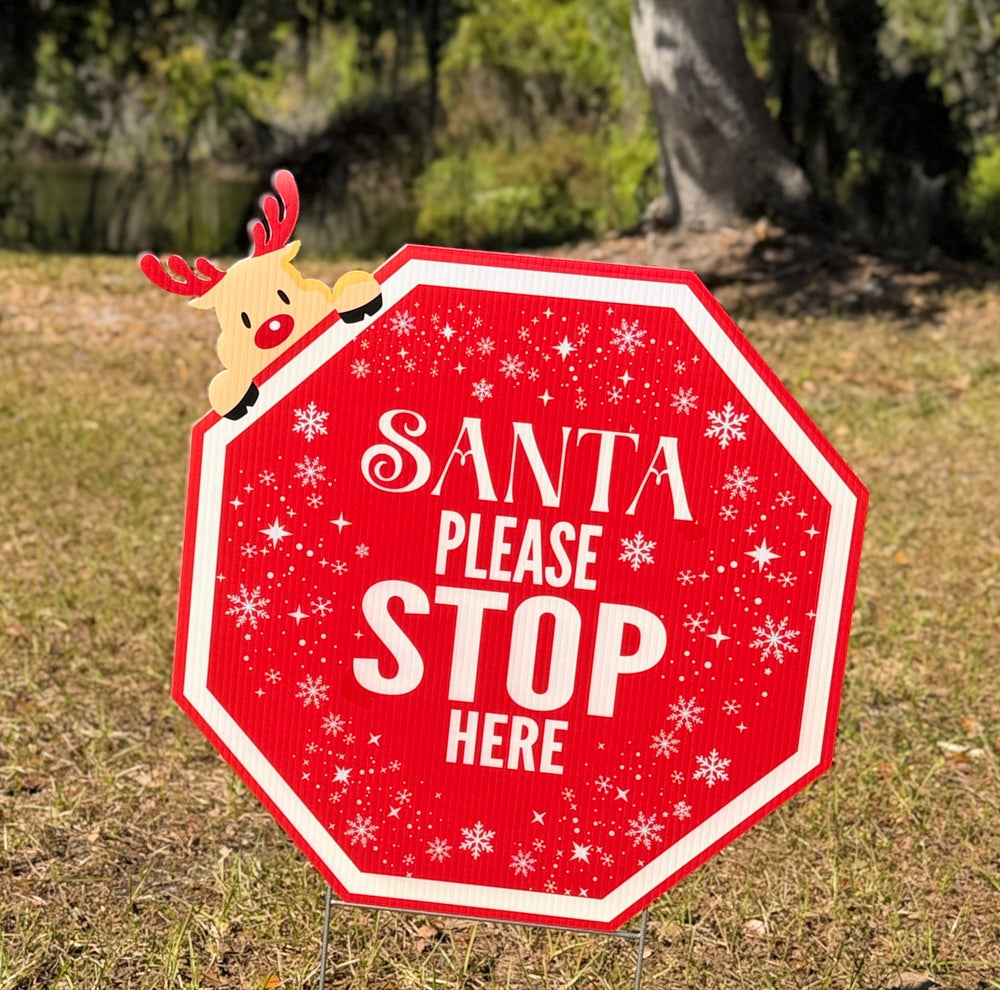 Red sign with 'Santa Please Stop Here' text and a reindeer figure on grass