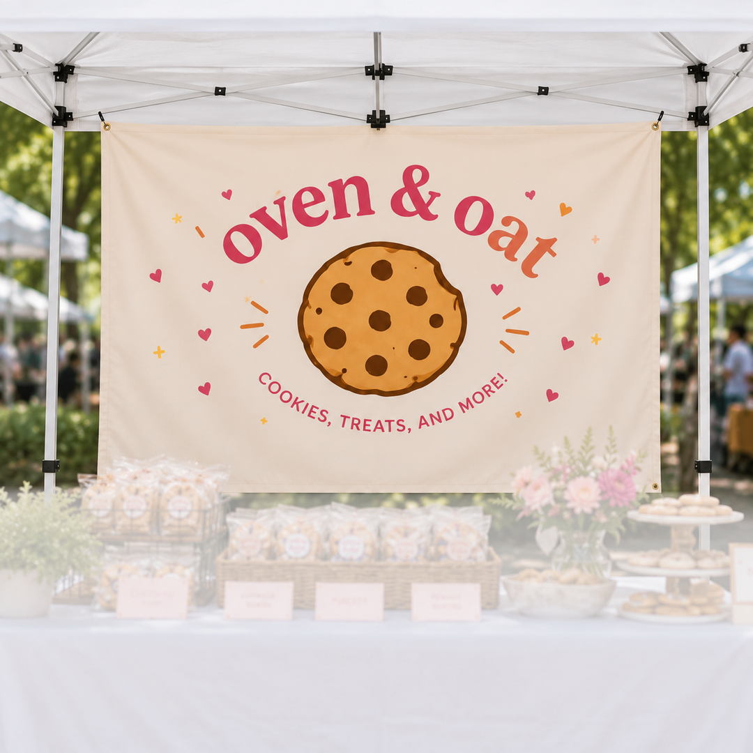 A cream-colored banner hanging under a white market canopy, featuring the words 'oven & oat' in coral-red script around a large chocolate chip cookie illustration. Smaller text reads 'COOKIES, TREATS, AND MORE!' with small pink hearts and orange/yellow sparkles.