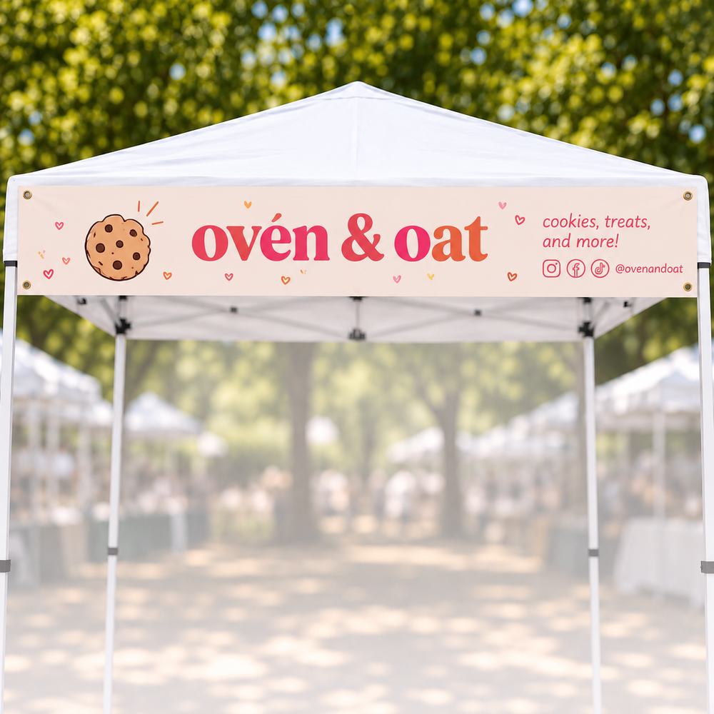 A white pop-up canopy tent with a printed banner reading “ovén & oat” and “cookies, treats, and more!” in a real outdoor market setting with blurred trees and other tents in the background.